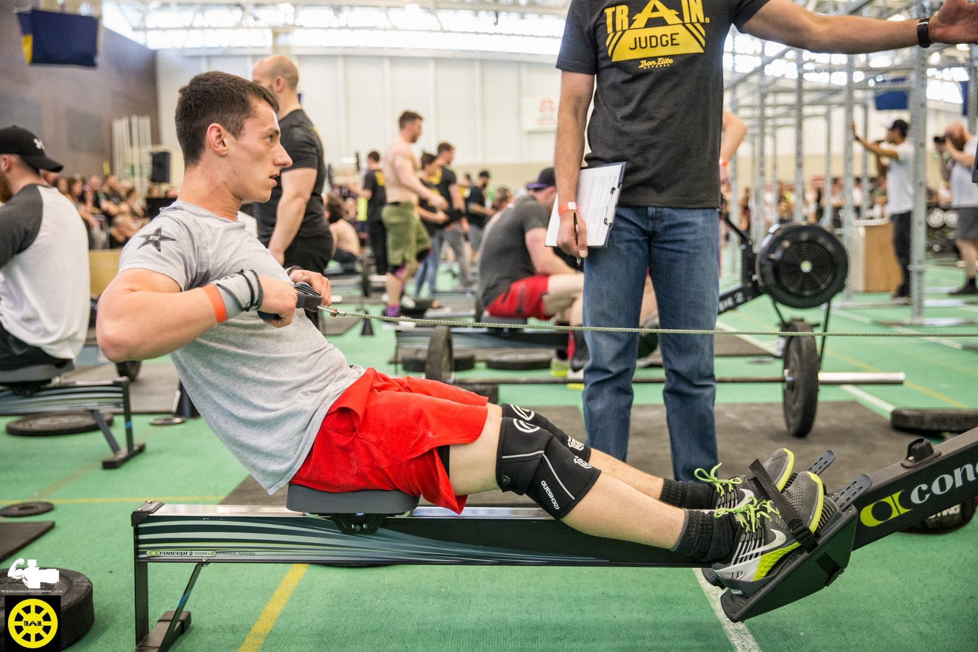 Nick mid-row on a Concept2 at a CrossFit competition, judge standing beside him
